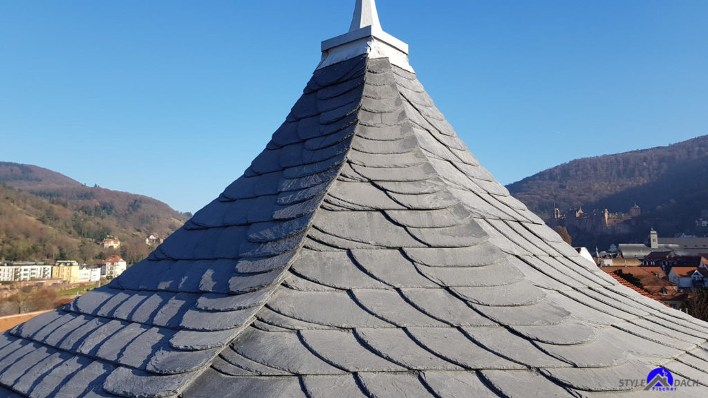 Das Bild zeigt einen Zwiebelturm der mit Schiefer Schuppen eingedeckt ist. Im Hintergrund sieht man den Neckar und die Stadt Heidelberg